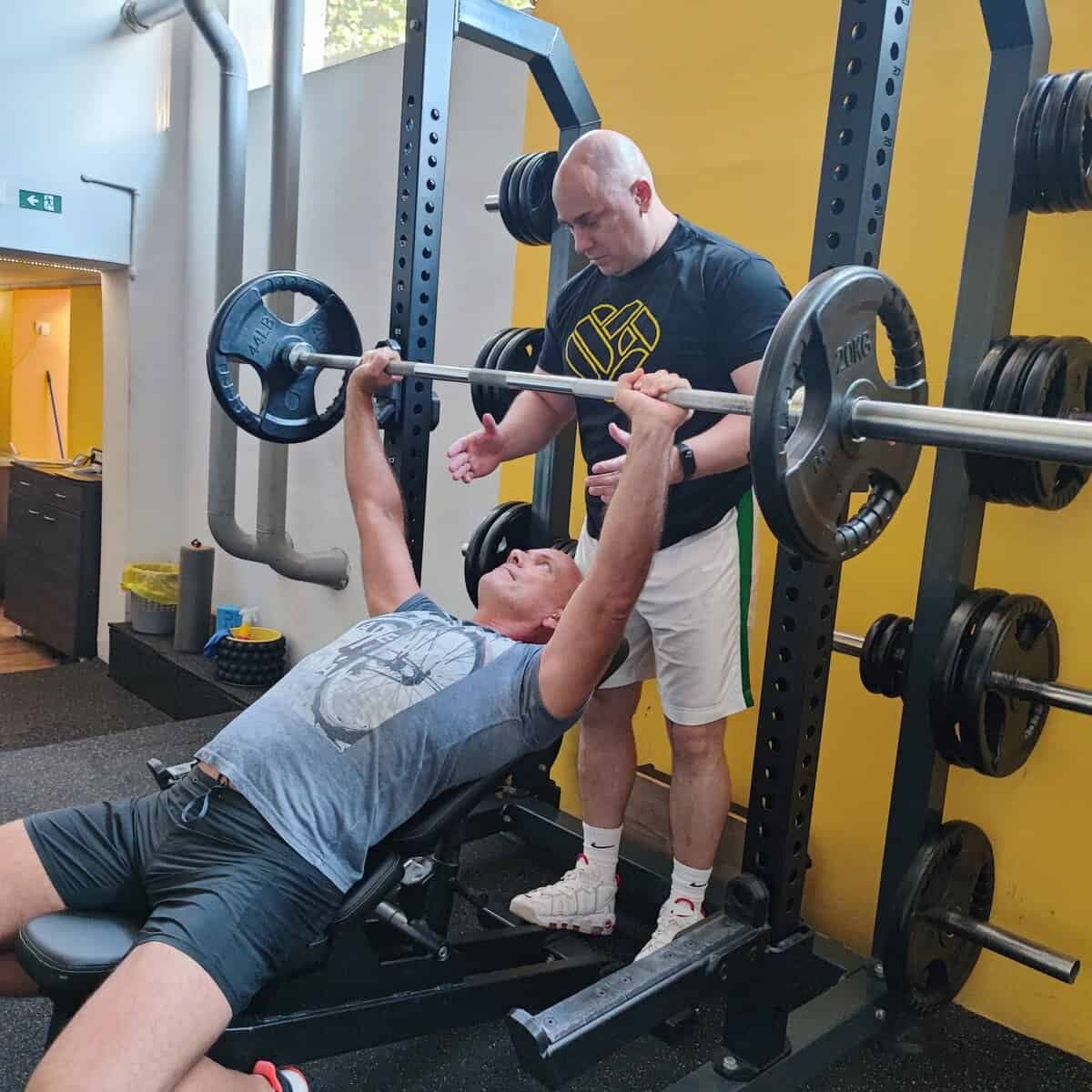 Trener asistira vežbaču pri vežbi sa šipkom na klupi u studiju Workout Team